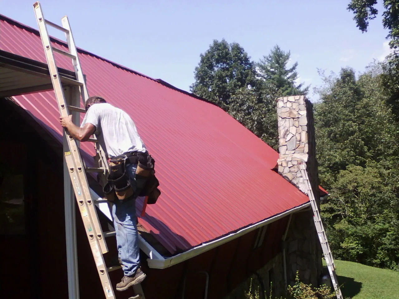 Roofer on a ladder beside a red metal roof and stone chimney — Soffit & Fascia Repair by Roofing Brothers in Fresno